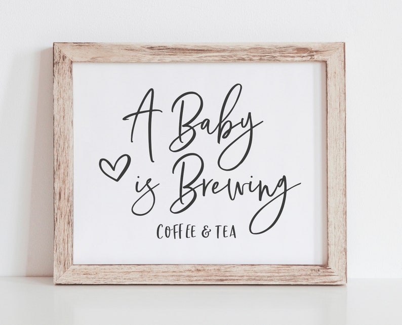 May include: A framed sign with the text "A Baby is Brewing Coffee & Tea" in black cursive script with a small heart above the word "is".
