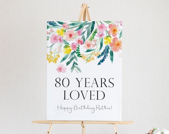May include: A white sign with a watercolor floral design in shades of pink, yellow, and blue. The sign says "80 Years Loved" and "Happy Birthday Ruthie!"