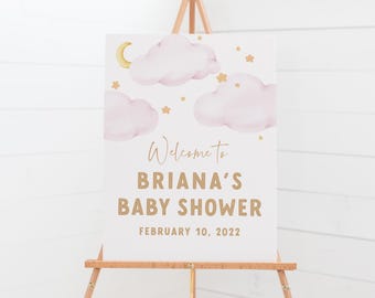 May include: A white sign on a wooden easel with the text "Welcome to Briana's Baby Shower February 10, 2022." The sign features watercolor illustrations of pink clouds, gold stars, and a crescent moon.