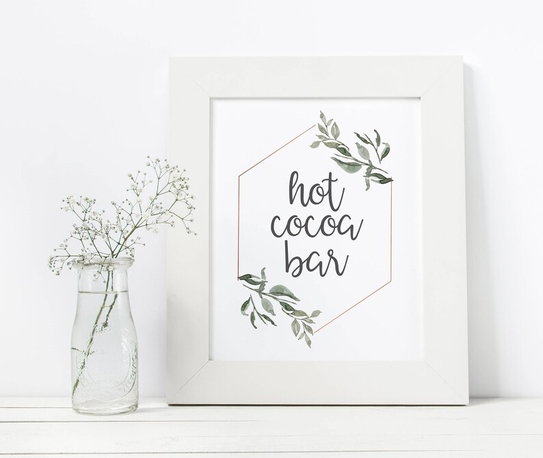 May include: White framed sign with a gold hexagon and green watercolor leaves. The text inside the hexagon reads "hot cocoa bar".