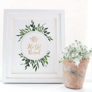 May include: A white framed print with a gold bee and the text "Let's Get Buzzed" surrounded by green watercolor leaves.