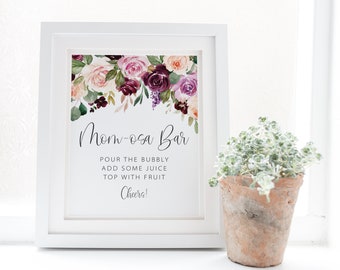 May include: A white framed sign with a floral design at the top. The sign says "Mom-osa Bar" and includes instructions for making a mimosa: "Pour the bubbly, add some juice, top with fruit. Cheers!"