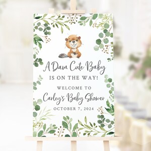 Könnte beinhalten: Ein weißes Schild mit einem grünen Blatt-Rand und einem niedlichen Cartoon-Biber. Das Schild sagt "A Dam Cute Baby is on the way! Welcome to Carley's Baby Shower October 7, 2024".