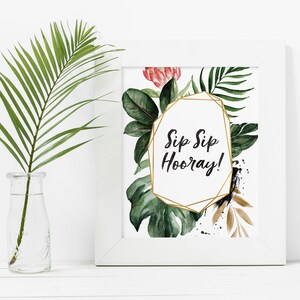 May include: A white framed print with a gold geometric frame and the text "Sip Sip Hooray!" surrounded by watercolor illustrations of tropical leaves and flowers.