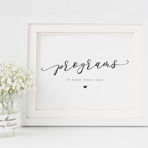 Könnte beinhalten: Weißer Rahmen mit einem Schild, auf dem "Programme bitte nehmen Sie eins" steht, darunter ein kleines schwarzes Herz. Das Schild steht auf einer weißen Oberfläche mit einem Glas voller weißer Blumen davor.