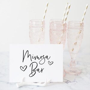 Peut inclure: Une pancarte blanche avec une calligraphie noire qui dit "Mimosa Bar" avec deux petits c&oelig;urs de chaque c&ocirc;t&eacute; du texte. La pancarte est pos&eacute;e sur une branche d'&eacute;lan blanche. Trois fl&ucirc;tes &agrave; champagne en verre rose avec des pailles dor&eacute;es sont en arri&egrave;re-plan.