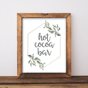 May include: A printable sign with a gold hexagon frame and green watercolor leaves. The sign says "hot cocoa bar" in a handwritten font.
