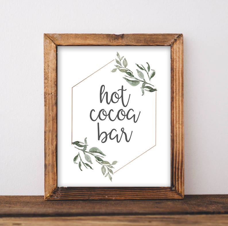 May include: A printable sign with a gold hexagon frame and green watercolor leaves. The sign says "hot cocoa bar" in a handwritten font.