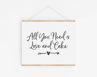 May include: A white sign with a wooden frame hanging on a white wall. The sign says "All You Need is Love and Cake" with a heart and arrows on either side of the text.