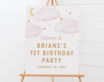 May include: Welcome to Briane's 1st Birthday Party sign with pink clouds, gold stars and a crescent moon. The sign is on an easel and the text is in gold.