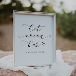 Pu&ograve; includere: Un cartello bianco con calligrafia nera che recita "hot cocoa bar" con un piccolo simbolo di cuore. Il testo "warm up here!" &egrave; stampato sotto la calligrafia.