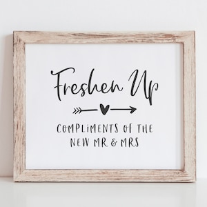 Può includere: Un cartello incorniciato con il testo "Freshen Up" con un design a freccia e cuore. Il testo "Compliments of the New Mr & Mrs" è sotto il design.