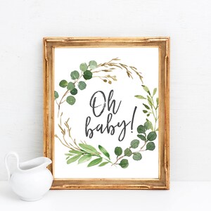 May include: A gold framed print with a white background and a green leafy wreath with the text "Oh baby!" in black.
