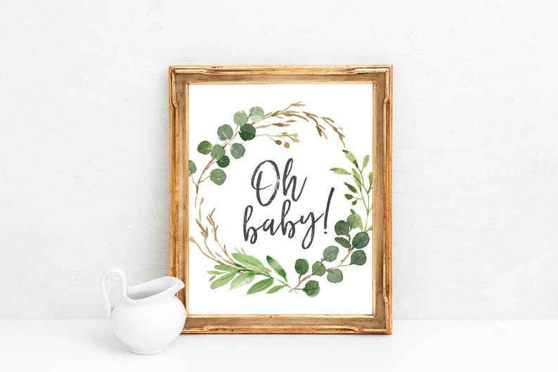 May include: A gold framed print with a white background and a green leafy wreath with the text "Oh baby!" in black.