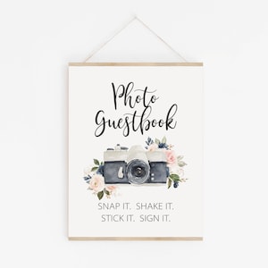 May include: A framed sign with the words "Photo Guestbook" in elegant script above a watercolor illustration of a camera surrounded by pink and blue flowers. The text "SNAP IT. SHAKE IT. STICK IT. SIGN IT." is below the camera.