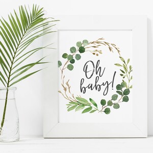 May include: A white framed print with a green and brown watercolor wreath with the text "Oh baby!" in black calligraphy.
