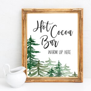 May include: A gold-framed sign with a watercolor illustration of a forest scene. The sign reads "Hot Cocoa Bar" and "Warm Up Here".