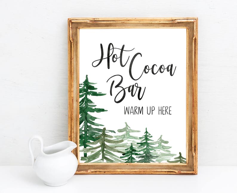 May include: A gold-framed sign with a watercolor illustration of a forest scene. The sign reads "Hot Cocoa Bar" and "Warm Up Here".