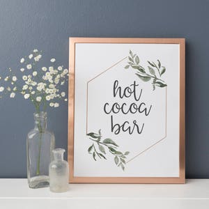 May include: A white sign with a copper frame and a gold hexagon design. The sign says "hot cocoa bar" with green watercolor leaves around the hexagon.