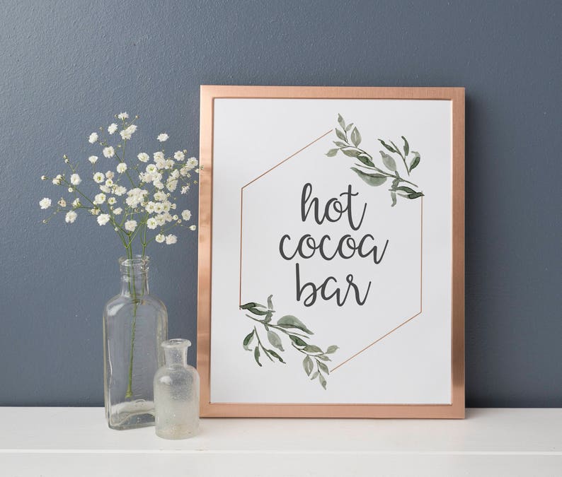 May include: A white sign with a copper frame and a gold hexagon design. The sign says "hot cocoa bar" with green watercolor leaves around the hexagon.