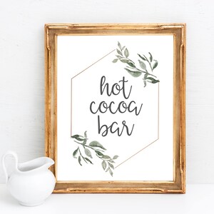 May include: A gold framed sign with a white background and a gold hexagon with green watercolor leaves. The text "hot cocoa bar" is written in black inside the hexagon.