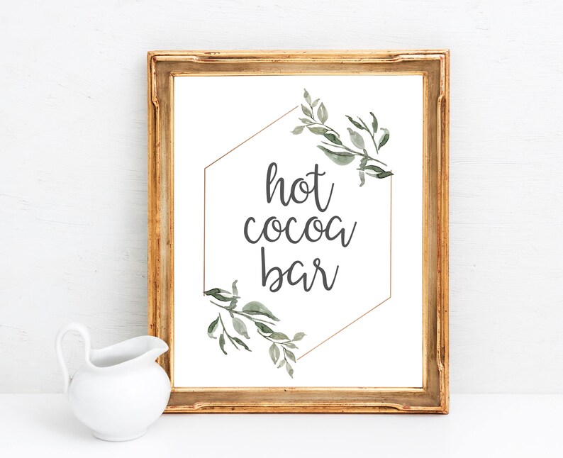 May include: A gold framed sign with a white background and a gold hexagon with green watercolor leaves. The text "hot cocoa bar" is written in black inside the hexagon.