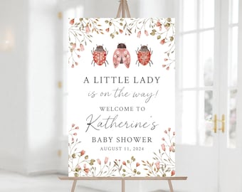 May include: A white sign with a floral design and three ladybugs. The sign reads "A little lady is on the way! Welcome to Katherine's Baby Shower August 11, 2024".
