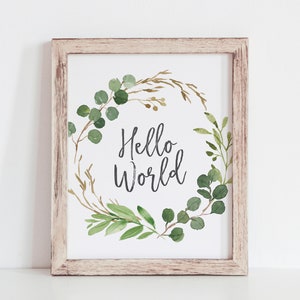 Op de afbeelding: Een ingelijst afdruk met een aquarel-illustratie van een groene eucalyptuskrans met de tekst "Hello World" in het midden.