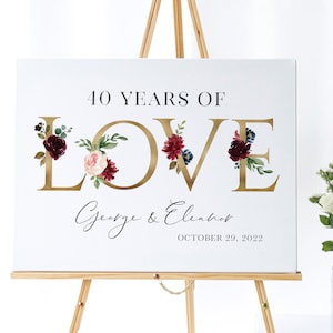 Könnte beinhalten: Ein weißes Schild mit goldenen Buchstaben, die "LOVE" buchstabieren, mit burgunderroten und rosafarbenen floralen Akzenten. Der Text "40 YEARS OF" befindet sich über dem Wort "LOVE" und der Text "George & Eleanor OCTOBER 29, 2022" befindet sich unter dem Wort "LOVE".