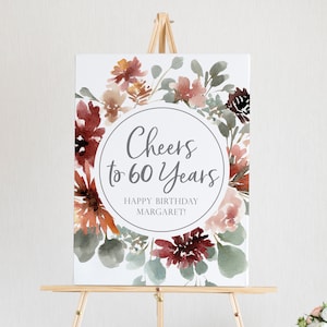 May include: A white sign with a watercolor floral design in shades of red, pink, and green. The sign says "Cheers to 60 Years" and "Happy Birthday Margaret!"