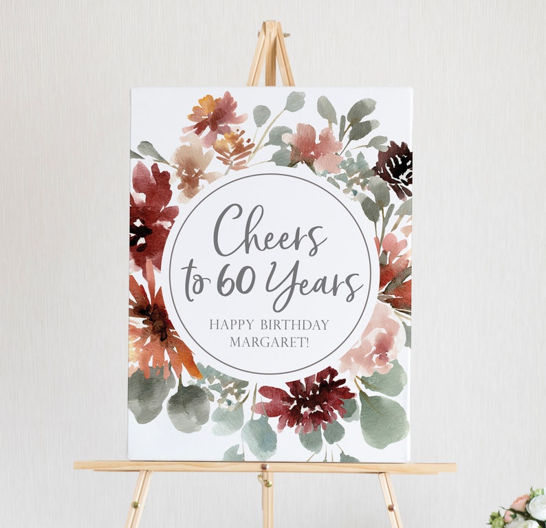 May include: A white sign with a watercolor floral design in shades of red, pink, and green. The sign says "Cheers to 60 Years" and "Happy Birthday Margaret!"