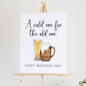 May include: A white sign on a wooden easel displays the text "A cold one for the old one" above watercolour illustrations of a beer glass and a beer mug. The text "HAPPY BIRTHDAY ERIK!" is below the illustrations.