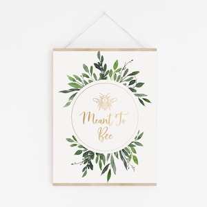 May include: A white printable sign with a gold geometric bee illustration and the text "Meant To Bee" in a gold script font. The bee and text are inside a gold circle surrounded by a wreath of green watercolor leaves.