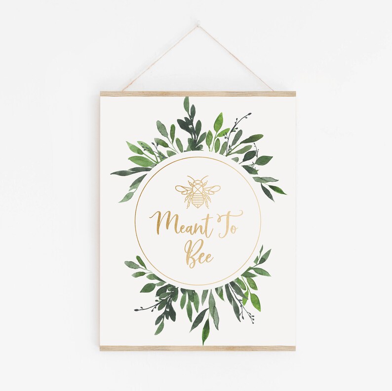 May include: A white printable sign with a gold geometric bee illustration and the text "Meant To Bee" in a gold script font. The bee and text are inside a gold circle surrounded by a wreath of green watercolor leaves.