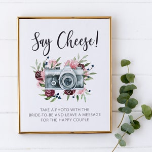 May include: A white sign with a gold frame and the text "Say Cheese!" in black script. A watercolour illustration of a vintage camera with pink and burgundy flowers surrounds the camera. The text "Take a photo with the bride-to-be and leave a message for the happy couple" is below the camera.