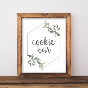 May include: A printable sign with a gold hexagon frame and green foliage. The sign reads "cookie bar" in a gray script font.
