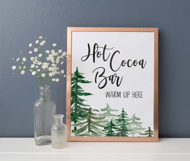 May include: A printable sign with a copper frame and a watercolor illustration of pine trees. The sign reads "Hot Cocoa Bar Warm Up Here".