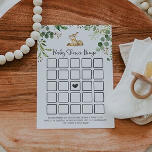 May include: A printable baby shower bingo card with a green and white floral design and a cute deer illustration. The card has a grid of squares with a heart in the center. The text "Baby Shower Bingo" is at the top of the card.