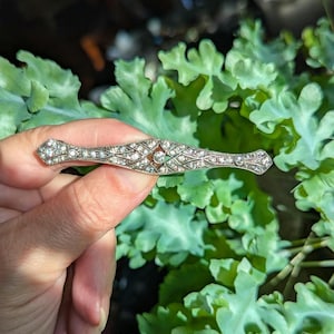 Puede incluir: Un broche de barra en tono plateado con un diseño geométrico de celosía alargada. La superficie metálica está incrustada con numerosas piedras pequeñas, claras y brillantes, con una piedra redonda central ligeramente más grande.