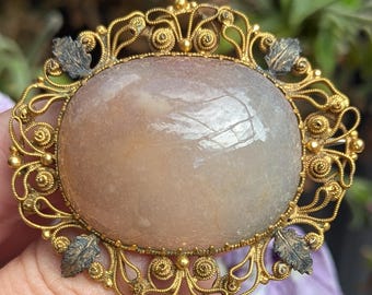 May include: An oval brooch featuring a large, smooth, translucent pale peach-colored stone set within an intricate gold-tone metal frame. The frame displays detailed filigree scrollwork and four dark, oxidized metal leaf accents at the cardinal points.