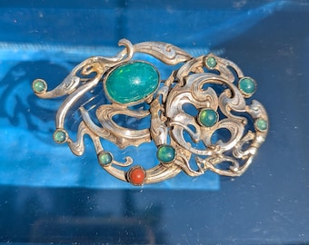 May include: A silver-toned metal brooch featuring an intricate, swirling openwork design. The piece is adorned with one large oval green cabochon, several smaller round green cabochons, and a single round orange cabochon set within the metal frame.