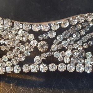Op de afbeelding: Een zilverkleurige haarspeld versierd met talrijke heldere, ronde strass-steentjes. De haarspeld heeft een bloemmotief met clusters van strass-steentjes en sierlijke, gebogen lijnen. De algemene vorm is langwerpig en licht gebogen.