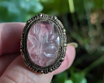 May include: An oval ring featuring a translucent pale pink carved gemstone with a floral motif. The stone is set in a dark, textured metal frame with a rope-like border and small spherical accents around the outer edge of the setting.