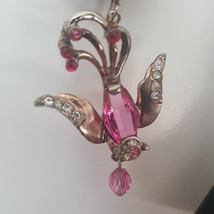 May include: A silver and pink bird-shaped earring with a pink crystal pendant. The earring is adorned with clear and pink rhinestones.