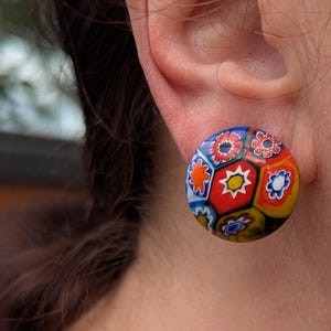 Könnte beinhalten: Runde Ohrstecker mit einem farbenfrohen Mosaikdesign. Die Ohrringe zeigen ein Muster aus roten, blauen, gelben und weißen Blumen- und geometrischen Formen. Ein einzigartiges Accessoire.