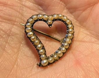 Puede incluir: Un broche en forma de corazón que presenta una serie de pequeñas perlas redondas de color crema dispuestas a lo largo de todo el perímetro del marco. Un fino mecanismo de alfiler metálico se extiende horizontalmente por el centro.