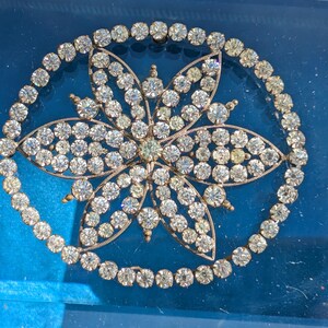 Puede incluir: Un broche de forma ovalada que presenta un diseño central de flor de seis pétalos incrustada con brillantes piedras de estrás transparentes. La flor está rodeada por un marco circular continuo de piedras de estrás a juego, montadas sobre una base metálica.