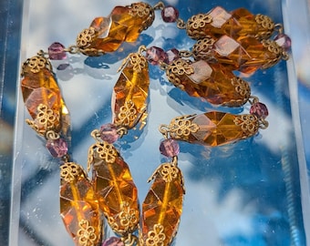 May include: A necklace featuring elongated, faceted amber-colored glass beads alternating with smaller, round purple beads. Each amber bead is capped at both ends with intricate, gold-toned metal filigree floral findings connected by metal links.