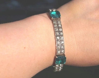 May include: A silver-toned bracelet featuring two large, rectangular, emerald-green stones at the ends and a central band composed of two parallel rows of small, clear, round, faceted stones set in a square metal frame pattern.