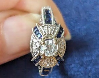 May include: A silver ring with a large, round, clear center stone surrounded by smaller clear stones and blue stones. The ring has a detailed design with a decorative band.